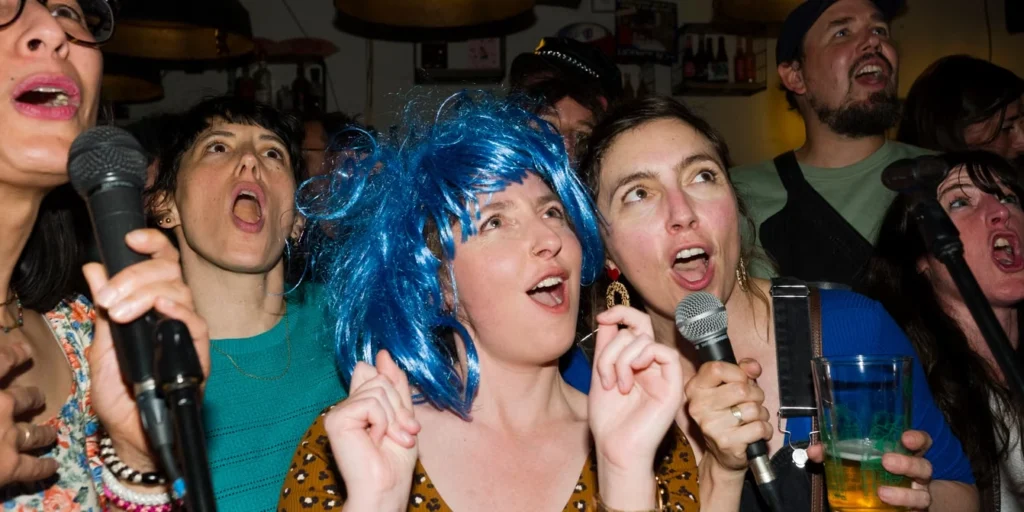 Groupe d’amis chantant au micro pendant une soirée karaoké, une invitée portant une perruque bleue